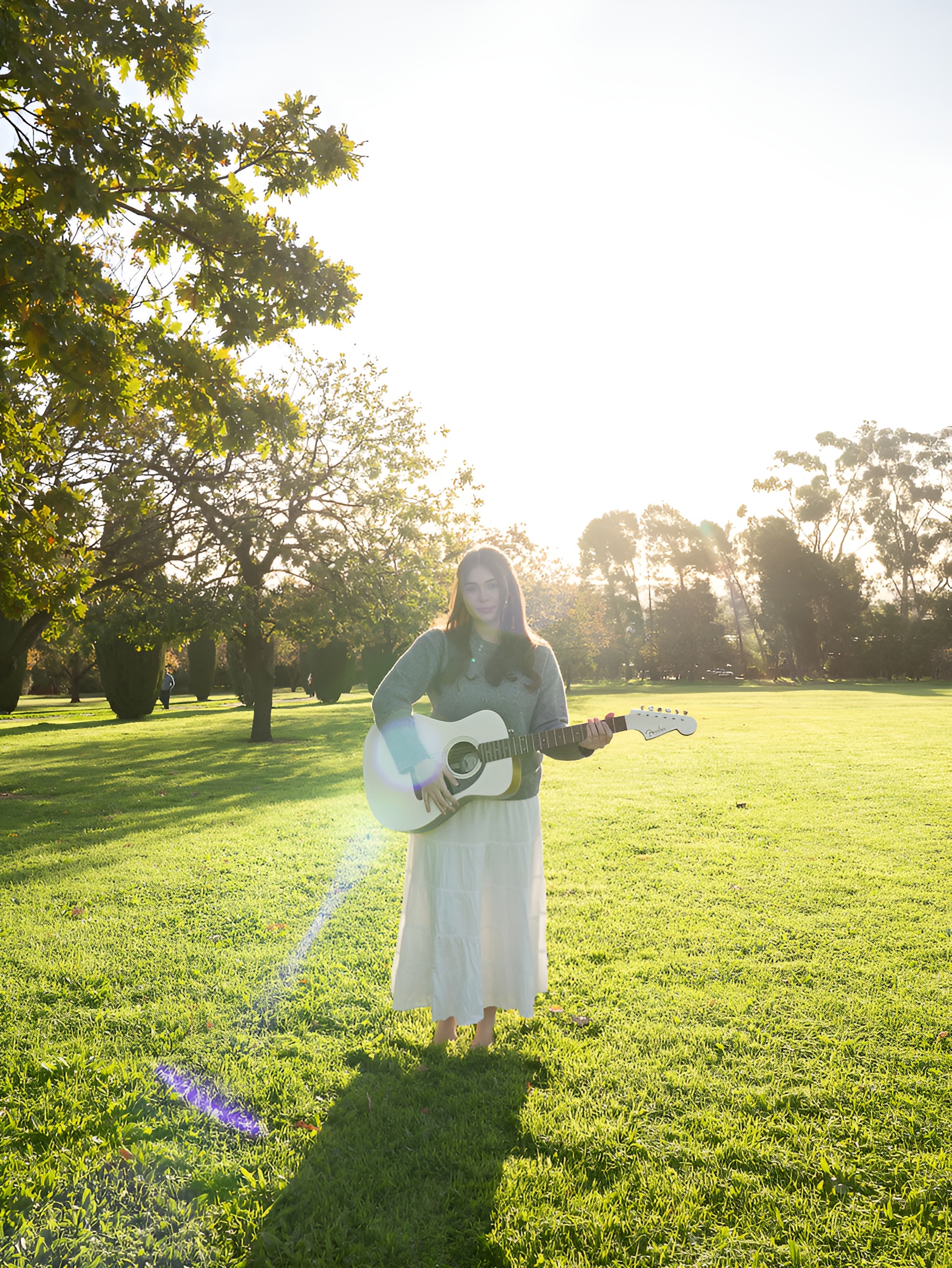 Ellise Hill standing in a sunlit field holding an acoustic guitar